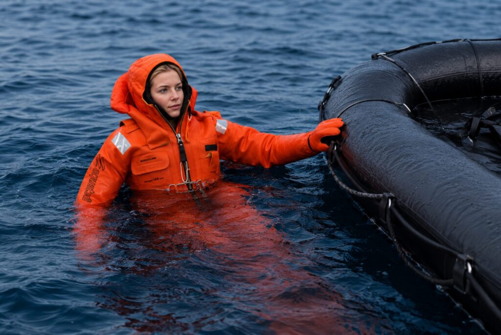Woman in an orange aquata immersion suit in the water next to a life raft
